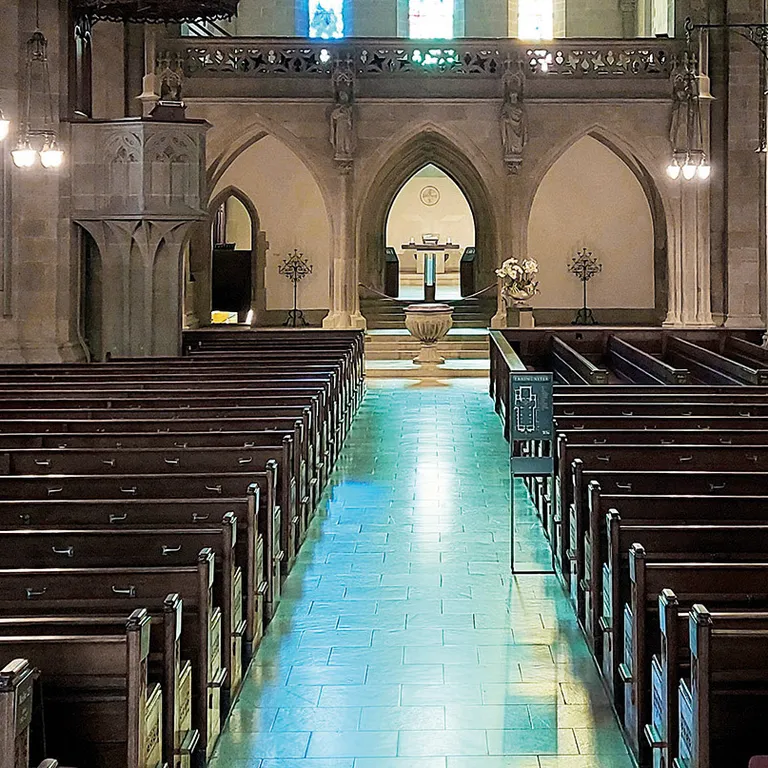 Innenraum einer Kirche mit Steinbögen, leeren Holzbänken und bunten Fenstern am Altar.