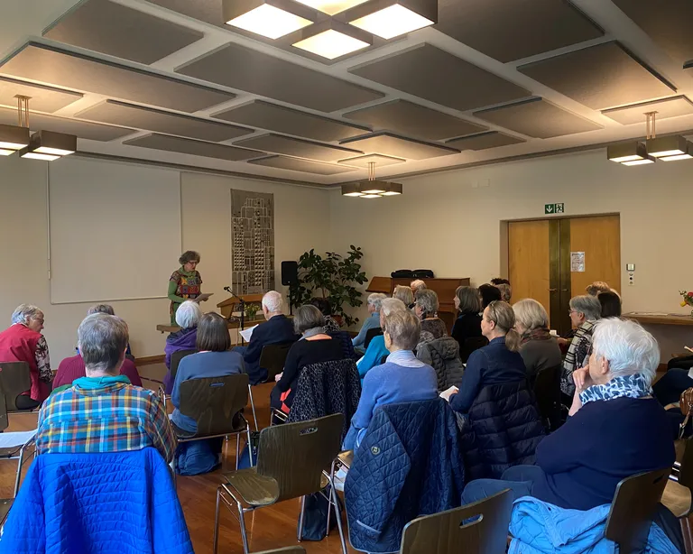 Mehrere Personen sitzen in einem Saal und hören einer stehenden Person vorne zu, die spricht oder präsentiert.