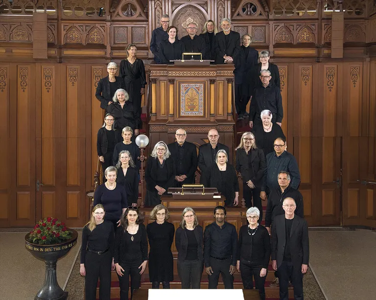 Eine größere Chorgruppe steht gestaffelt in einer Kirche vor einer Orgel, alle tragen dunkle Kleidung.