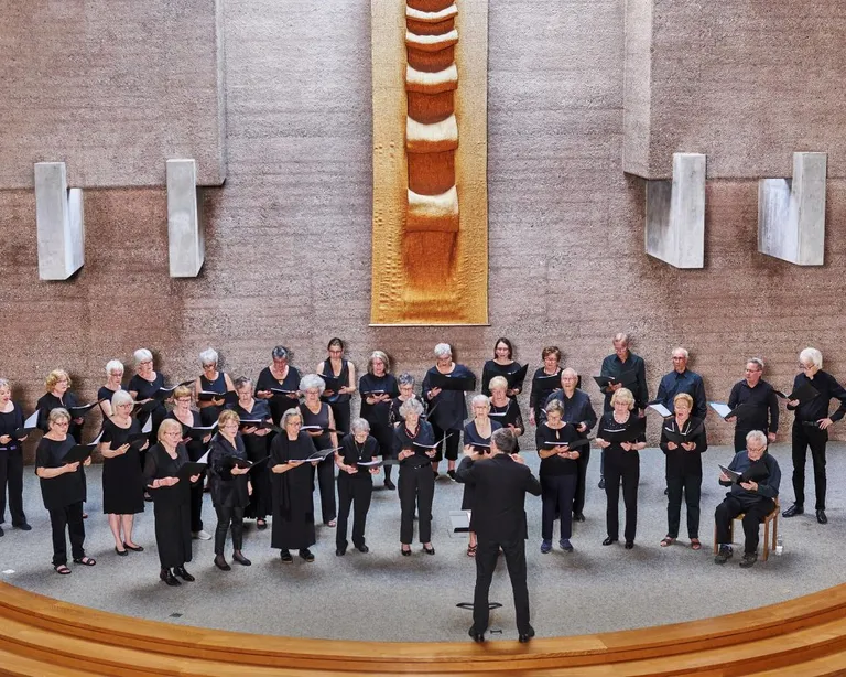 Ein Chor steht in einer modernen Kirche und singt, der Dirigent steht vorne und leitet die Gruppe.