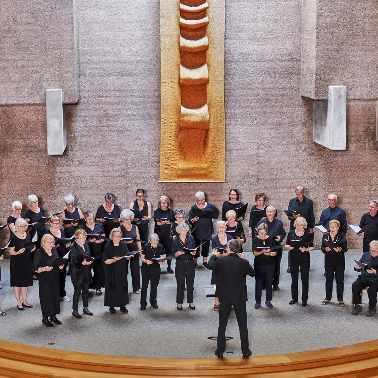 Ein Chor in schwarzer Kleidung singt in einer Kirche, dirigiert von einer Person, im Hintergrund moderne Kirchenarchitektur.