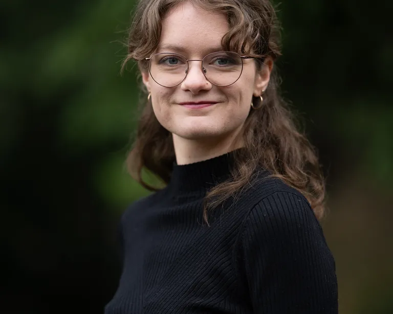Eine junge Frau mit Brille und lockigem Haar trägt einen schwarzen Pullover und steht im Freien vor unscharfem, grünem Hintergrund.