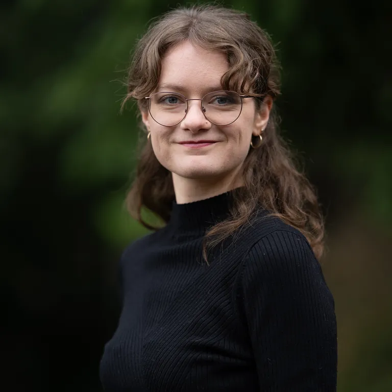 Eine junge Frau mit Brille und lockigem Haar trägt einen schwarzen Pullover und steht im Freien vor unscharfem, grünem Hintergrund.