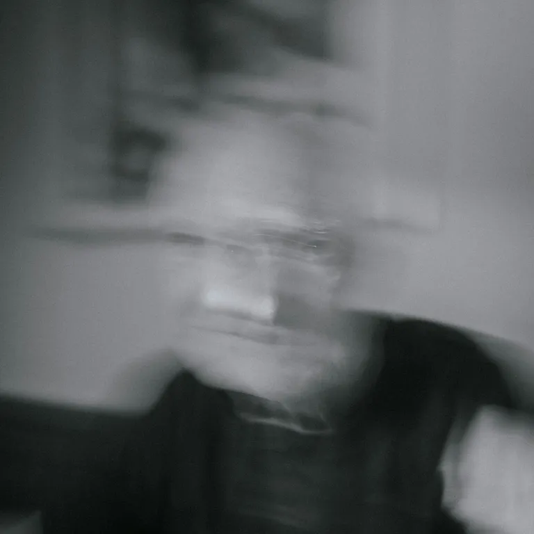 Verschwommenes Schwarz-Weiss-Foto einer sitzenden Person mit unklaren Gesichtszügen in einem Innenraum.
