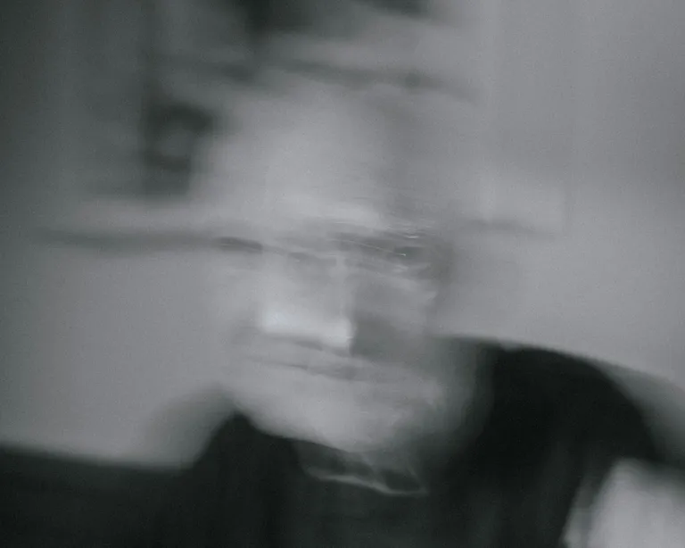 Verschwommenes Schwarz-Weiss-Foto einer sitzenden Person mit unklaren Gesichtszügen in einem Innenraum.