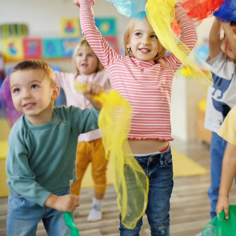 Vier fröhliche Kinder spielen in einem Raum und halten bunte Tücher in den Händen.