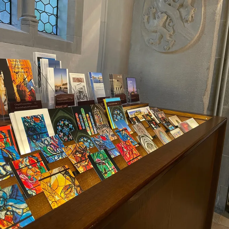 Bücher und Postkarten mit bunten Motiven sind auf einem Holztisch vor einem Fenster in einem hellen Raum ausgestellt.
