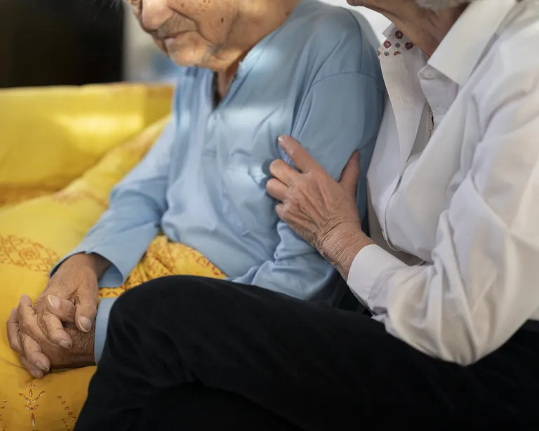 Eine Person pflegt eine ältere Person, beide sitzen eng beieinander auf einem gelben Sofa.
