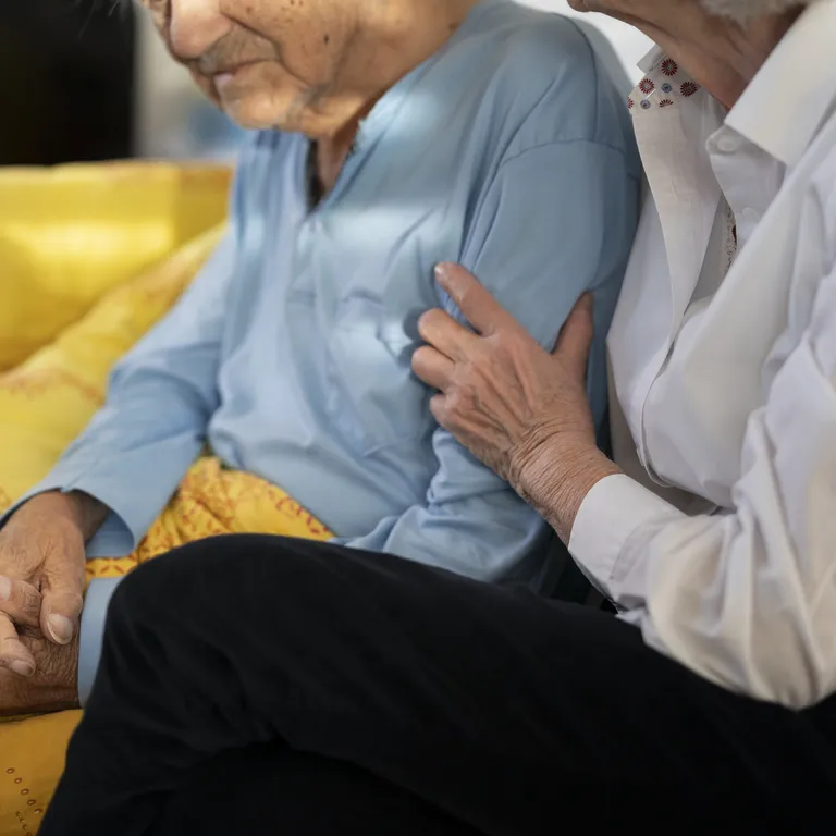 Eine Person pflegt eine ältere Person, beide sitzen eng beieinander auf einem gelben Sofa.