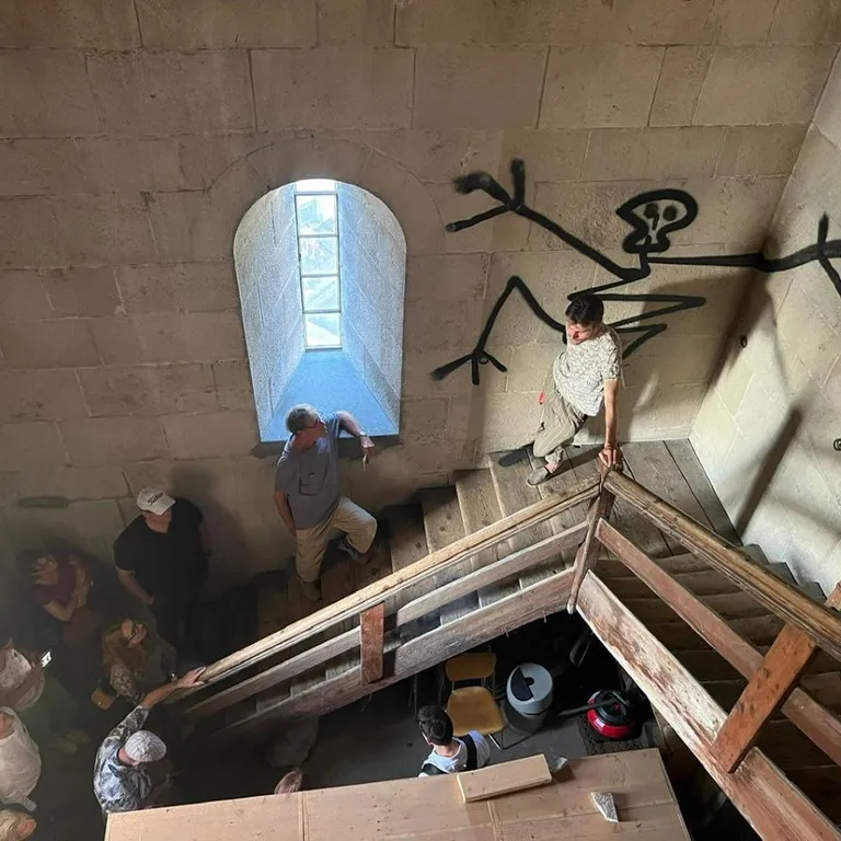 Eine kleine Gruppe steht in einem engen Treppenhaus in einem Kirchturm mit Graffiti-Wandbild in Form eines Strichmännchens und kleinem Fenster