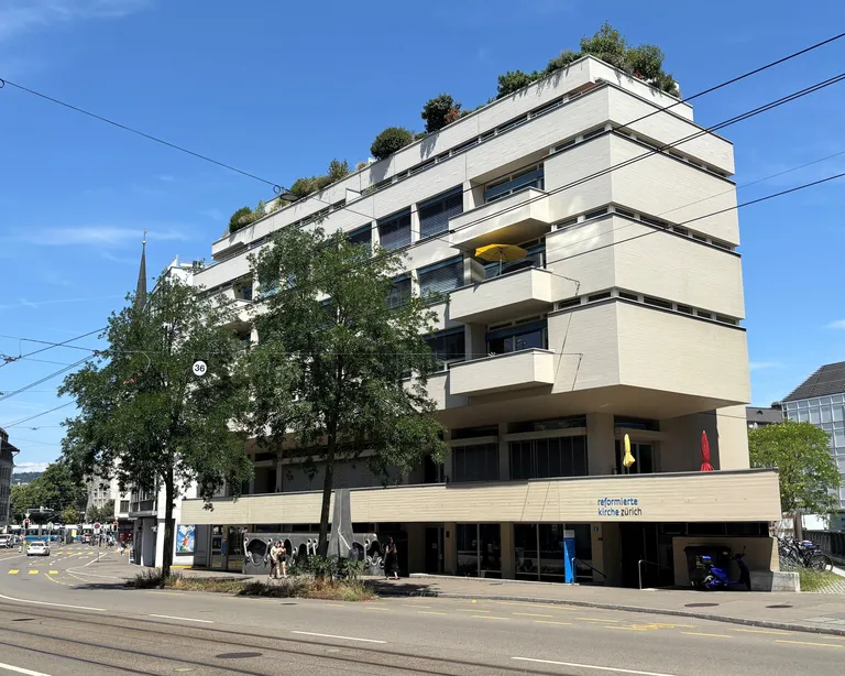 Ein mehrstöckiges Gebäude mit Balkonen an einer Strasse, Fahrradständern und einem Baum im Vordergrund.