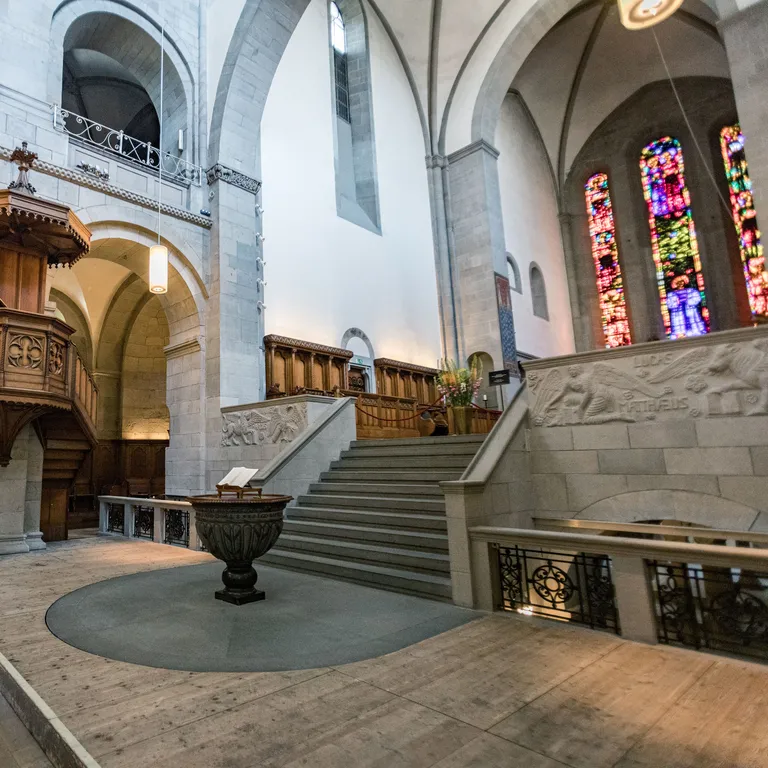 Heller Kircheninnenraum mit Taufbecken, Kanzel, Treppe zum Hochchor und bunten Glasfenstern der Künstler Sigmar Polke und Augusto Giacometti.