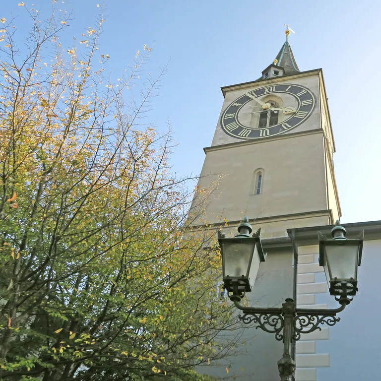 Kirchturm mit grosser Uhr, daneben eine Strassenlaterne und ein Baum, blauer Himmel im Hintergrund.