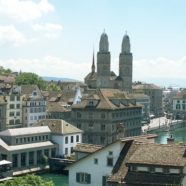 A view of Zurich featuring the Grossmünster, City Hall, other historic buildings, and the Limmat River under a blue sky.