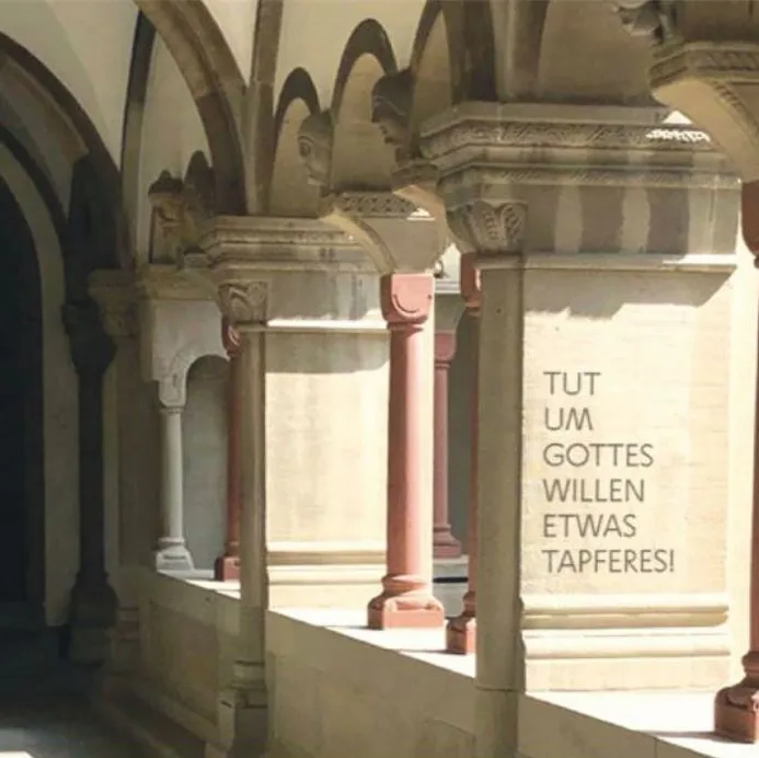 Ein Arkadengang mit Säulen und dem Schriftzug: «Tut um Gottes Willen etwas Tapferes».