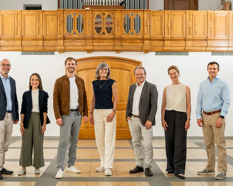Sieben Menschen stehen nebeneinander in einem hellen Raum mit Holzgalerie und Orgel im Hintergrund.