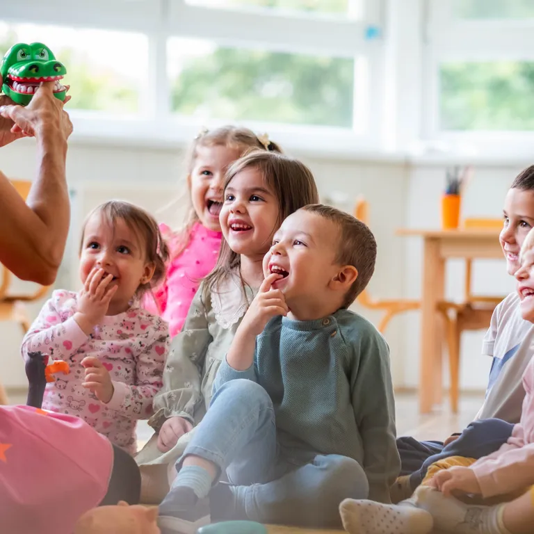 Mehrere Kinder sitzen lachend auf dem Boden und schauen gespannt einer Erwachsenen mit einer Handpuppe zu.