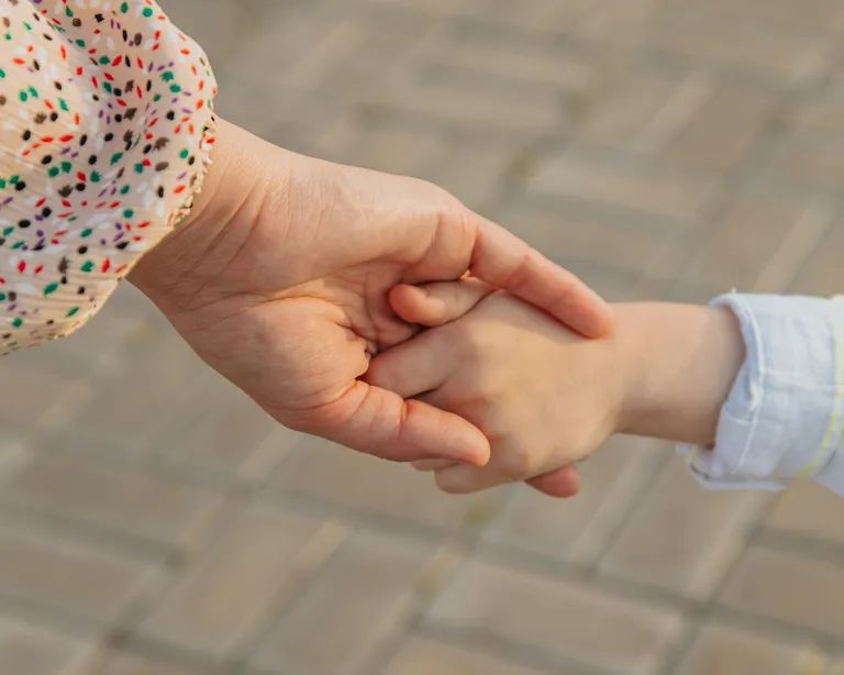 Eine erwachsene und eine Kinderhand halten sich auf einem gepflasterten Untergrund.
