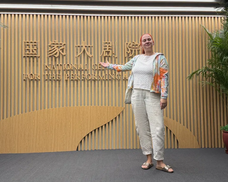 Eine Person steht lächelnd vor einer Holz-Wand mit chinesischer Schrift und grünen Pflanzen rechts und links.