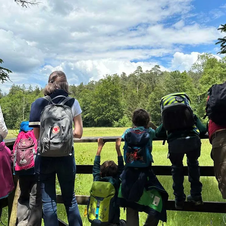Mehrere Menschen mit Rucksäcken stehen und sitzen an einem Zaun, blicken auf eine grüne Wiese unter blauem Himmel.