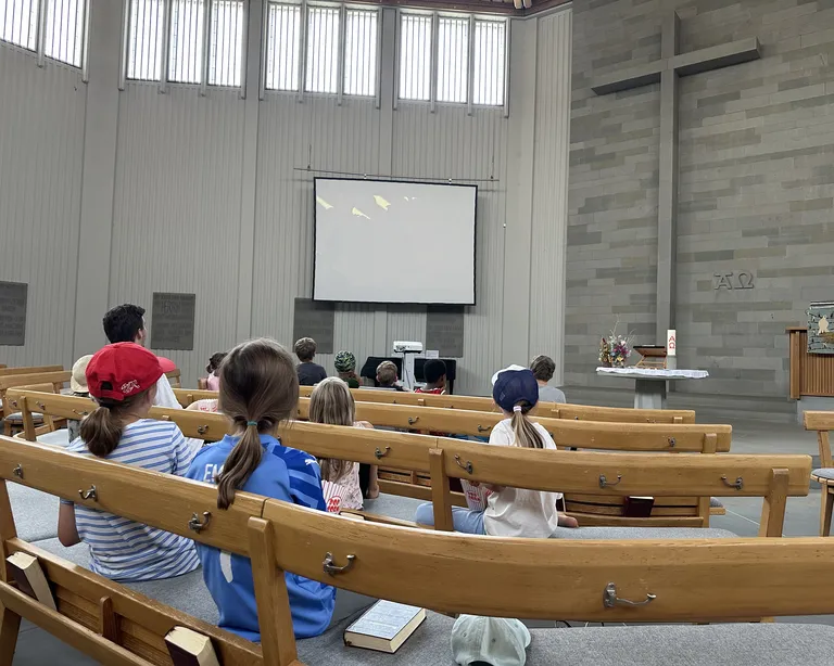 Kinder sitzen in einer Kirche auf Holzbänken, vor einem Altar mit großem Kreuz und einer Leinwand.