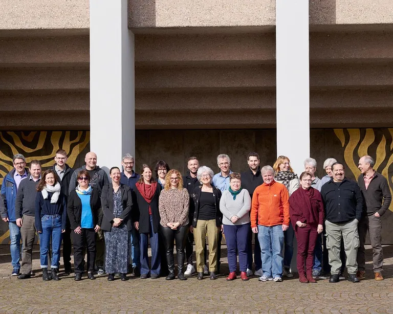 Eine Gruppe von etwa 25 Personen steht im Freien vor einem Gebäude mit gemusterter Wand und Säulen.
