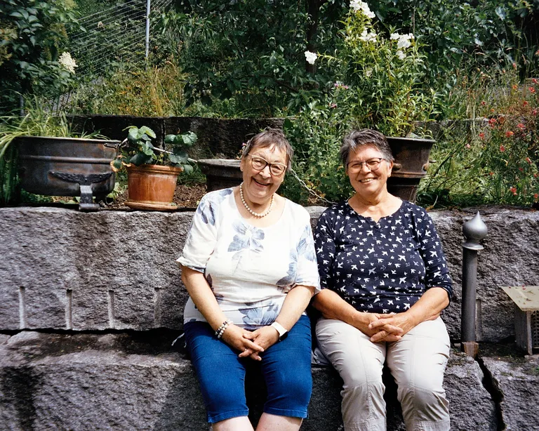 Zwei ältere Frauen sitzen nebeneinander auf einer Steinmauer im Garten und lächeln in die Kamera.