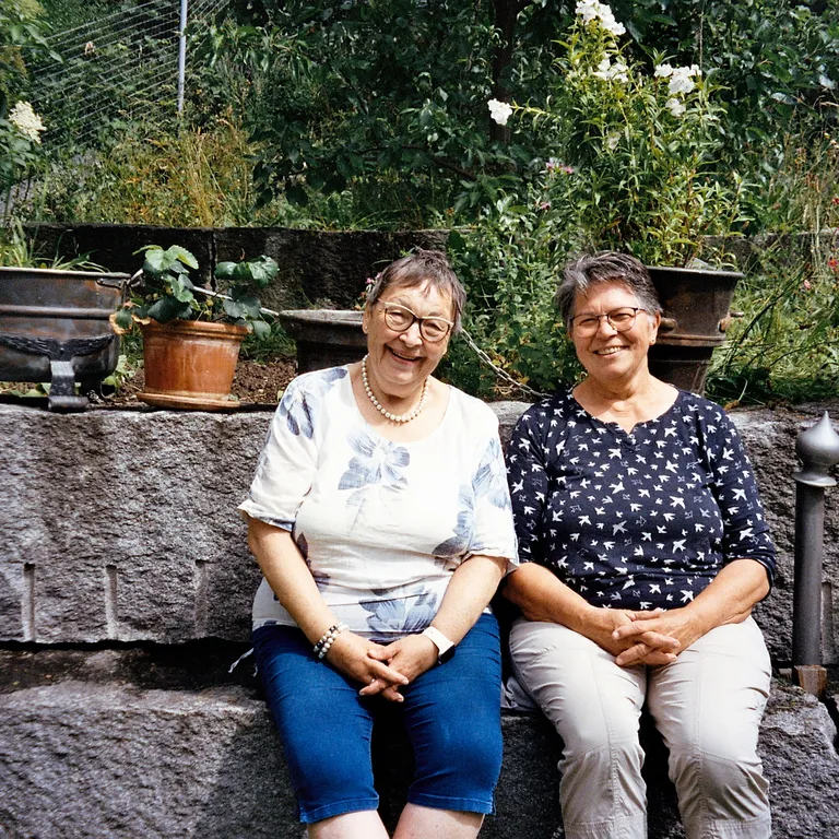 Zwei ältere Frauen sitzen nebeneinander auf einer Steinmauer im Garten und lächeln in die Kamera.