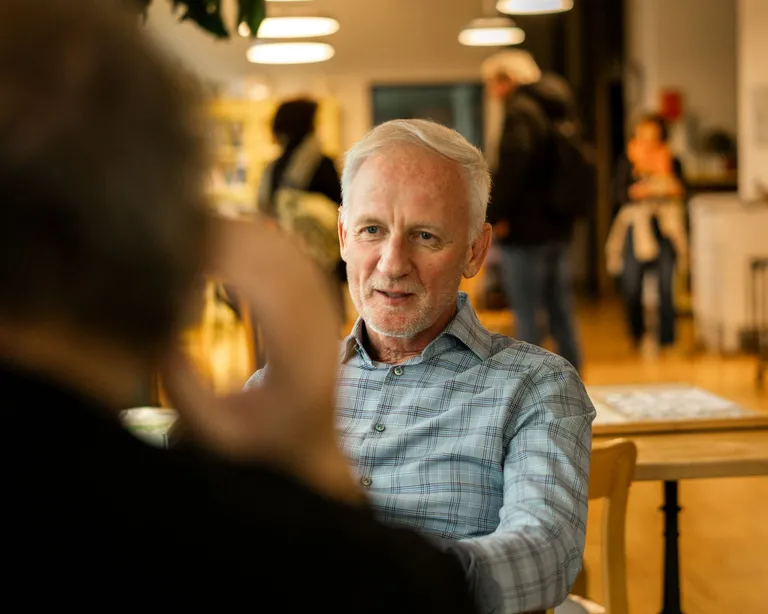 Ein älterer Mann sitzt in einem Café und unterhält sich mit einer weiteren, unscharfen Person im Vordergrund.