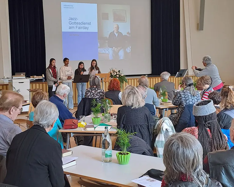 Menschen sitzen an Tischen in einem Saal, vorne sprechen vier Personen auf einer Bühne vor einer Leinwand.