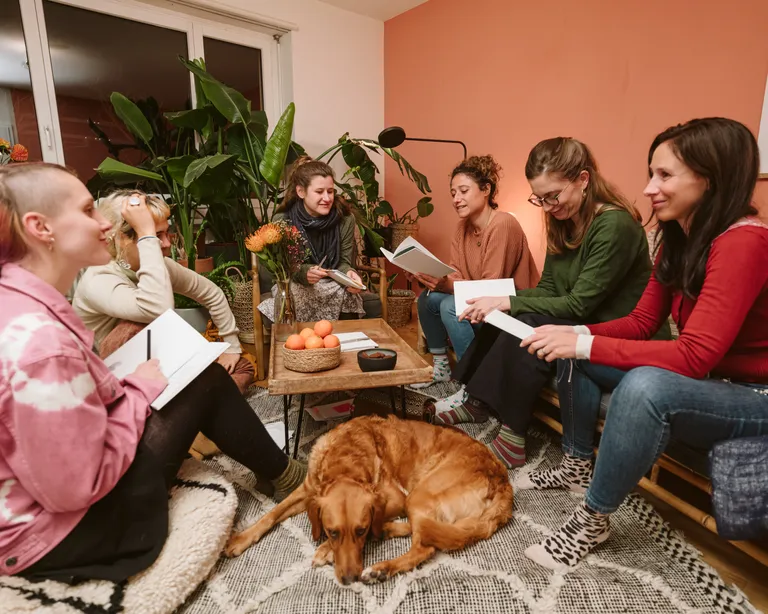Sechs Menschen sitzen lachend im Kreis in einem Wohnzimmer, lesen Bücher, ein Hund liegt auf dem Teppich.