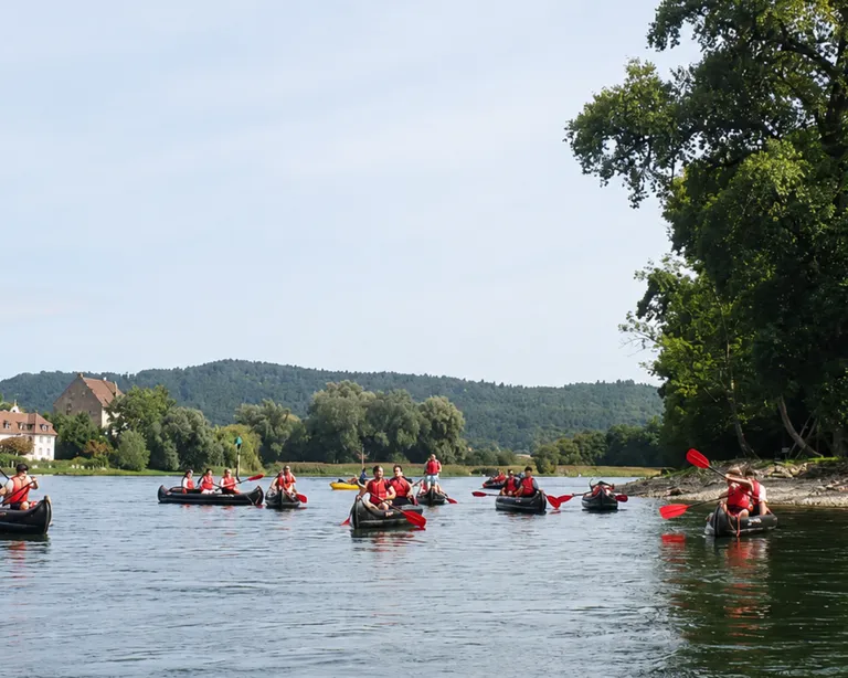 Mehrere Teilnehmende des Kurses für Jugendleiterinnen in Kanus auf einem ruhigen Fluss, im Hintergrund Bäume und Häuser, rechts ein grünes Ufer.