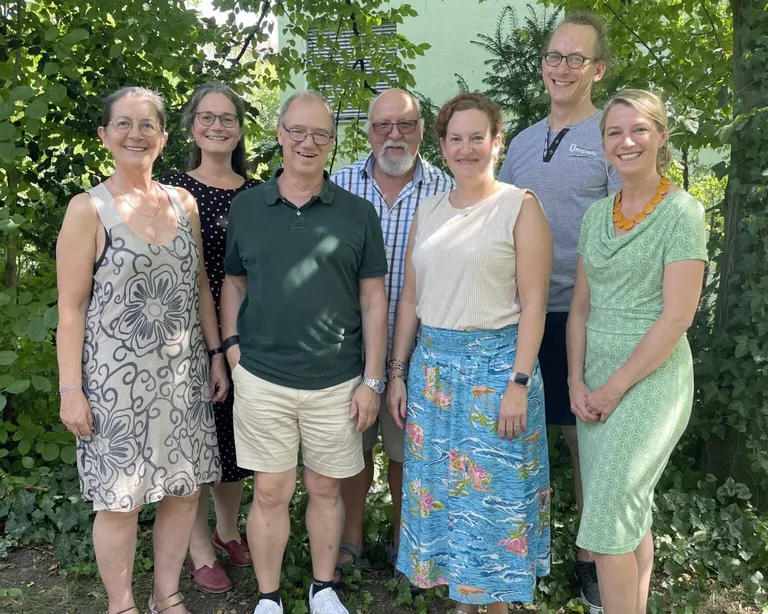 Sieben Mitglieder der Kirchenkreiskommission zehn stehen im Freien nebeneinander und lächeln in die Kamera. Im Hintergrund sind grüne Bäume.