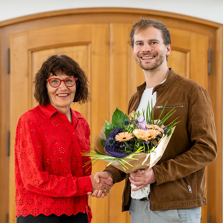 Eine Frau in roter Bluse und ein Mann mit Blumenstrauss geben sich vor einer Holztür die Hand und lächeln.