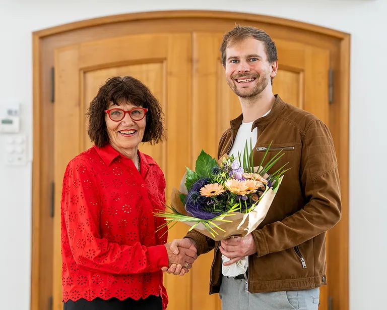 Eine Frau in roter Bluse und ein Mann mit Blumenstrauss geben sich vor einer Holztür die Hand und lächeln.