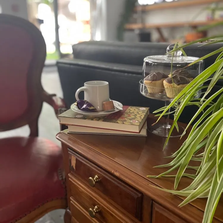 Holztisch mit Tasse, Büchern, Gebäck und einer Pflanze, im Hintergrund ein Sessel und ein Sofa.
