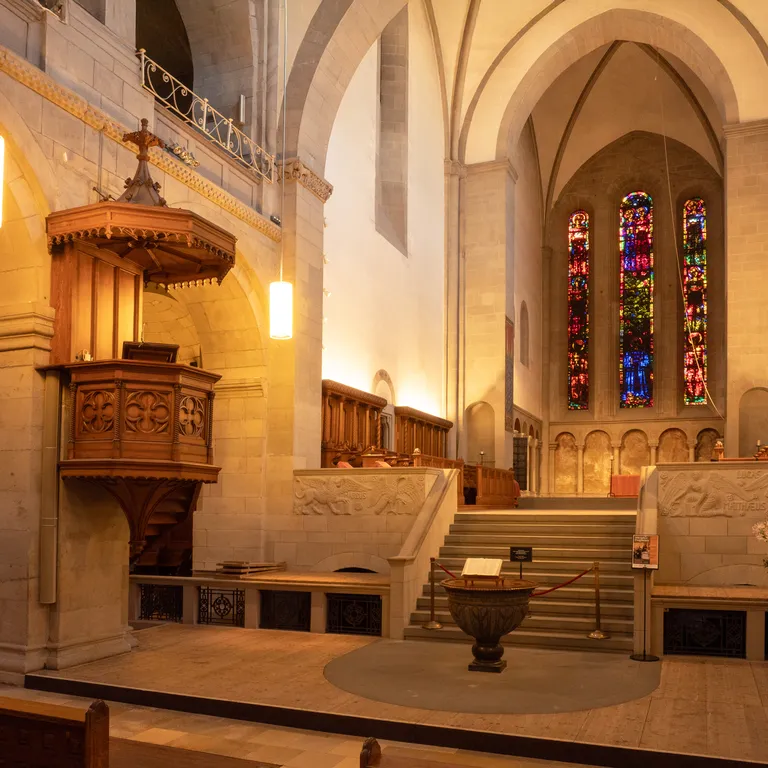 Innenraum der Kirche Grossmünster mit Kanzel, Taufbecken, Bänken und bunten Glasfenstern am Altar, stimmungsvolles Licht.