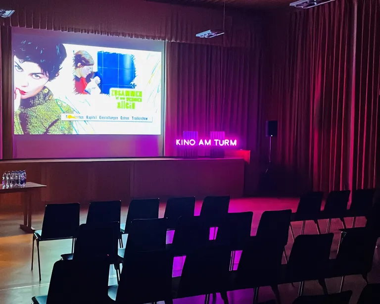 Kleiner Kinosaal mit Stuhlreihen, Leinwand mit Filmprojektion und pink leuchtendem «Kino am Fluss»-Schriftzug.