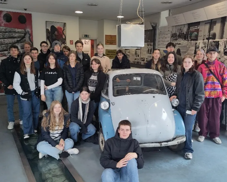 Eine Gruppe junger Menschen posiert vor einem kleinen, alten Auto in einem Museum.