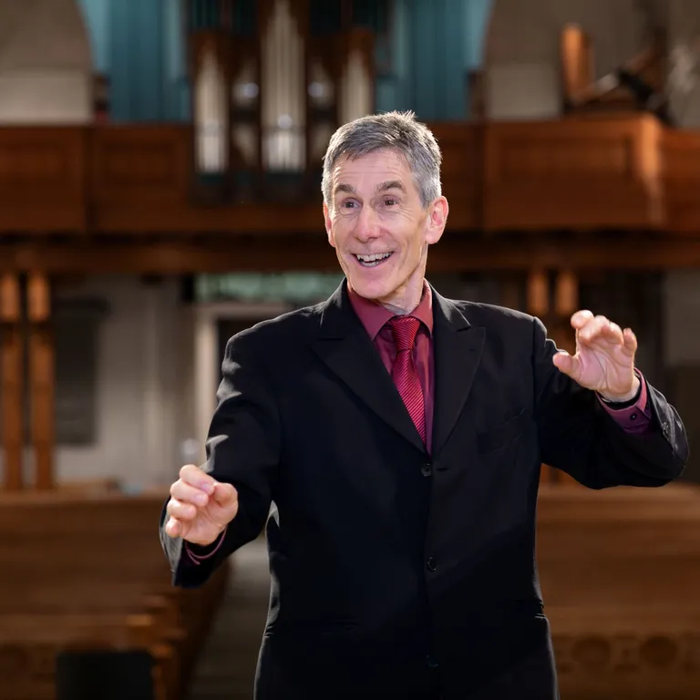 Ein Mann im Anzug dirigiert in einer Kirche, im Hintergrund sind Bänke und eine Orgel zu sehen.