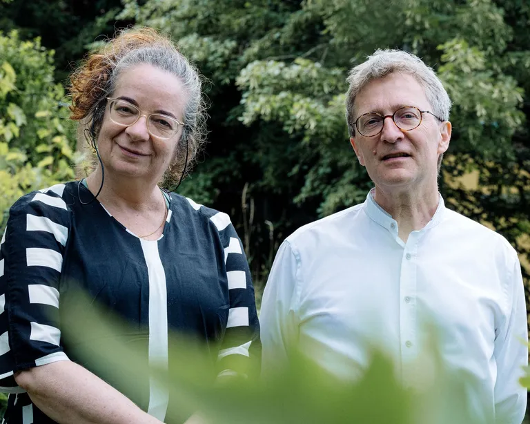 Zwei Menschen stehen nebeneinander im Garten vor grünen Bäumen und lächeln in die Kamera.