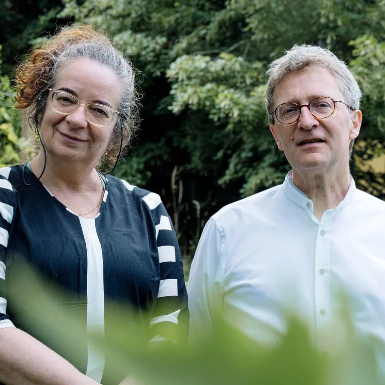 Zwei Menschen stehen nebeneinander im Garten vor grünen Bäumen und lächeln in die Kamera.