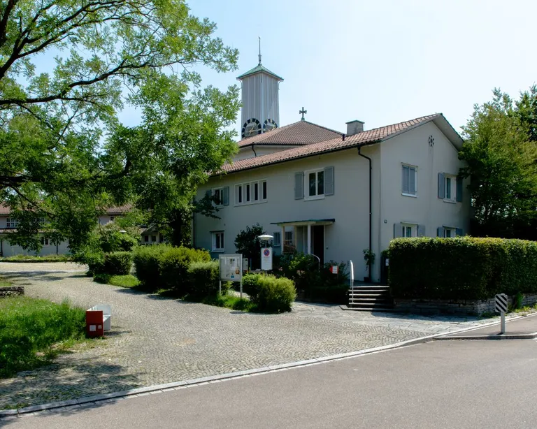 Ein zweistöckiges, helles Gebäude mit rotem Dach, umgeben von Bäumen und Büschen, im Vordergrund eine gepflasterte Einfahrt.
