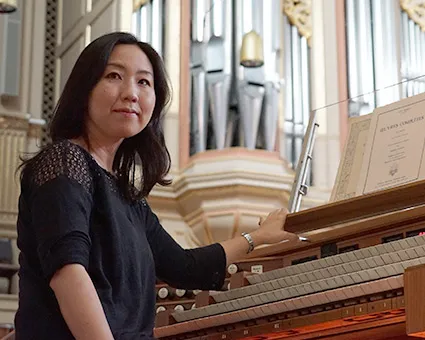 Frau sitzt an einer Orgel und blickt in die Kamera, im Hintergrund große Orgelpfeifen in einem Kirchenraum.