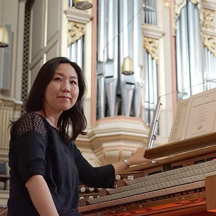 Frau sitzt an einer Orgel und blickt in die Kamera, im Hintergrund große Orgelpfeifen in einem Kirchenraum.