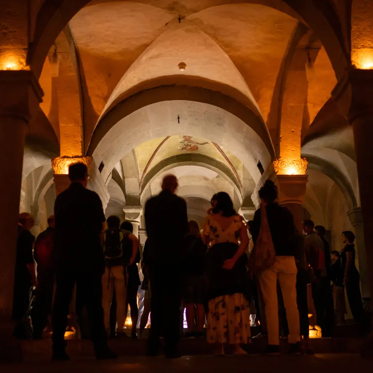 Mehrere Menschen stehen in einer stimmungsvoll mit Kerzen beleuchteten Halle mit gewölbter Decke und Bögen.