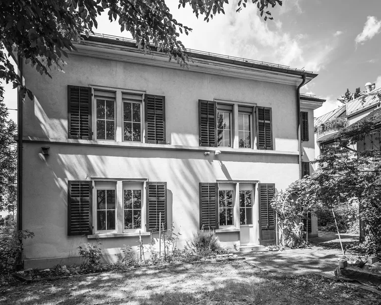 Schwarz-weiss-Foto des Pfarrhaus Wipkingen mit Fensterläden, Bäumen im Vordergrund und Schatten auf der Fassade.