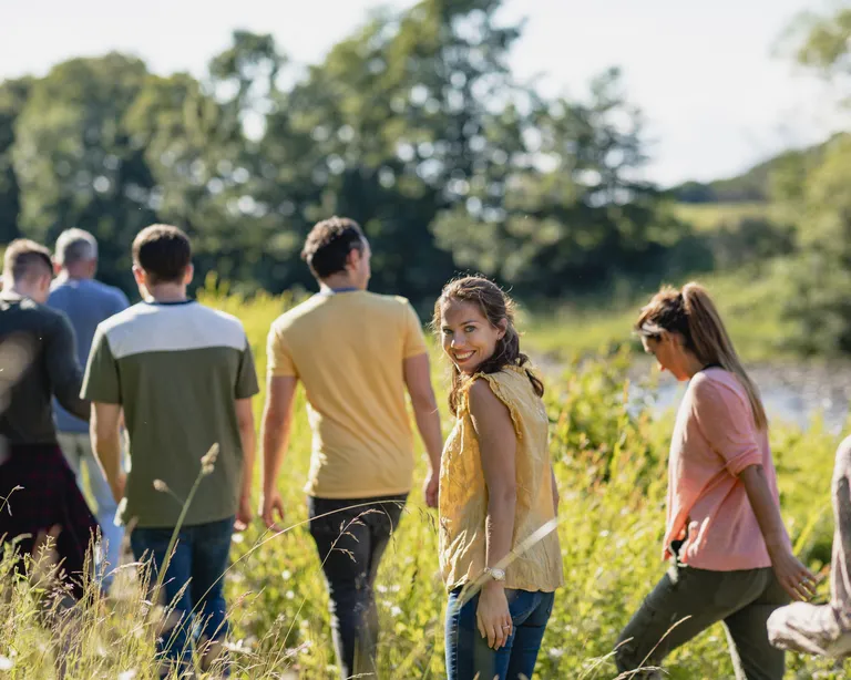 Eine Gruppe von Menschen spaziert durch eine grüne Wiese an einem sonnigen Tag, im Hintergrund Bäume und blauer Himmel.