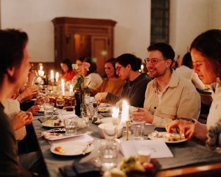 Mehrere Menschen sitzen an einem festlich gedeckten Tisch und essen gemeinsam bei Kerzenlicht.