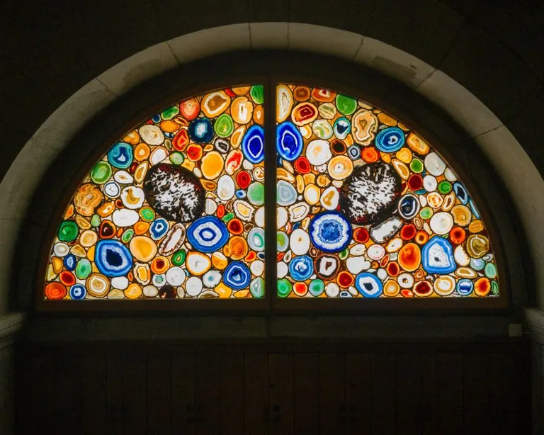 Halbrundes Fenster mit buntem Mosaikglas in Kreis- und Blumenmustern, beleuchtet vor dunklem Hintergrund.